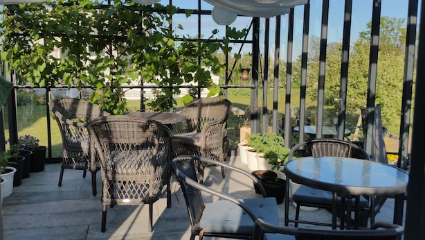 Eine überdachte Terrasse mit Korbmöbeln und einem Glastisch, umgeben von Blumentöpfen und Blick auf einen Garten.
