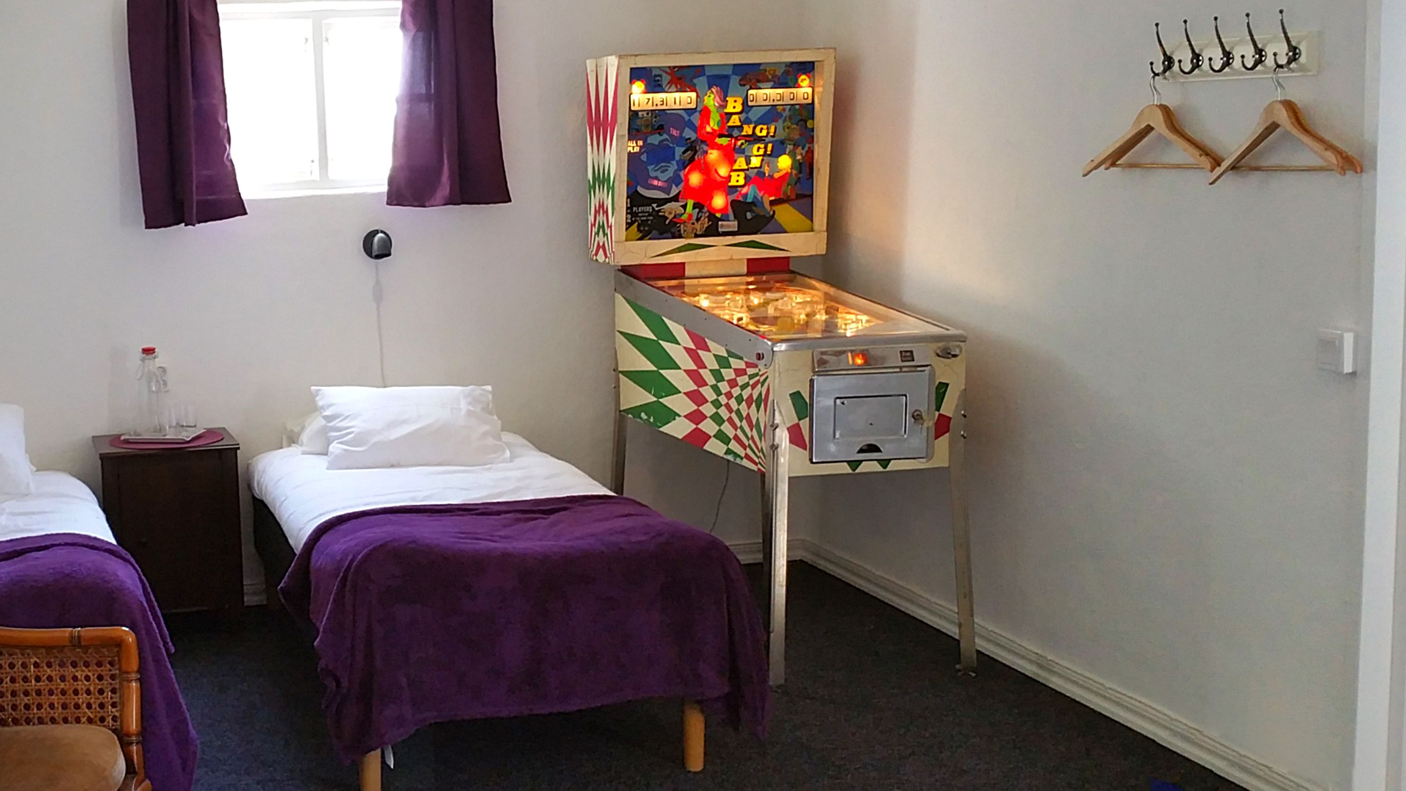 Une chambre d’hôtel avec deux lits simples, une petite table avec une bouteille et des verres, un flipper et un porte-manteau avec des cintres.