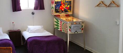 A hotel room with two single beds, a small table with a bottle and glasses, a pinball machine, and a coat rack with hangers.