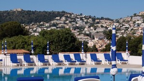 A pool area with blue lounge chairs and umbrellas, overlooking a hillside with buildings.