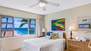 A hotel room with a large bed, a ceiling fan, a window with a beach view, a colorful painting, and a nightstand with towels. - Stunning Oceanfront, 2BR/2BA, Gorgeous Ocean & Mt. Views, Premium A/C (Kihei)