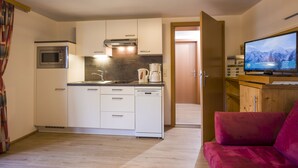 A compact kitchen with a microwave, stove, and dishwasher. A television is mounted on the wall, and a red sofa is in the living area.