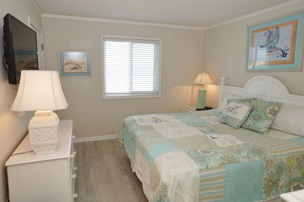 A bedroom with a bed, a nightstand, a lamp, a window, and two framed pictures on the wall. - Beachfront 3 Bedroom Condo at beautiful Ocean Isle Beach, NC (Ocean Isle Beach)