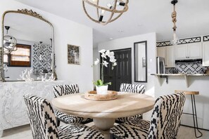 Un comedor con una mesa redonda, sillas estampadas, una araña y una cocina con armarios blancos.