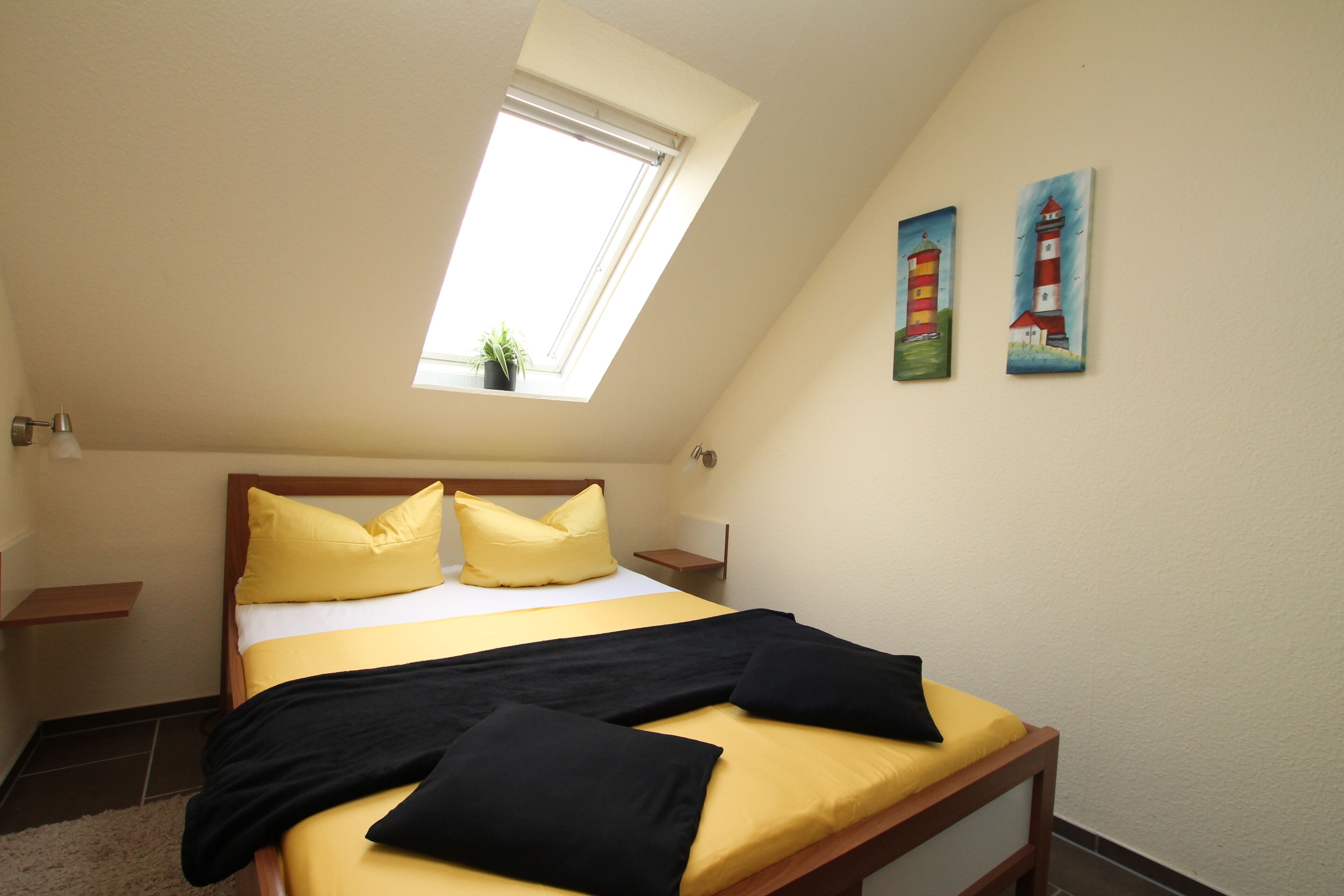A bedroom with a skylight, a bed with yellow and black bedding, two paintings on the wall, and a small plant on the windowsill.
