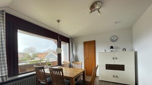 Comedor con mesa y sillas de madera, ventilador de techo, radiador y vistas a casas a través de amplios ventanales.