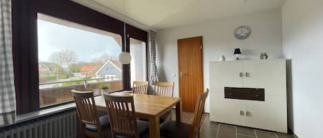 Ein Essbereich mit Holztisch und Stühlen, einem Deckenventilator, einem Heizkörper und Blick auf Häuser durch große Fenster.