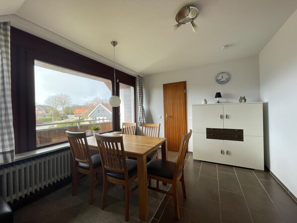 Un coin repas avec une table et des chaises en bois, un ventilateur de plafond, un radiateur et une vue sur les maisons à travers de grandes fenêtres.