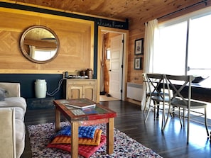 A cozy living room with a wooden ceiling, a round mirror, a small table with a book, a sofa, a dining area with chairs, and a door leading to another room.