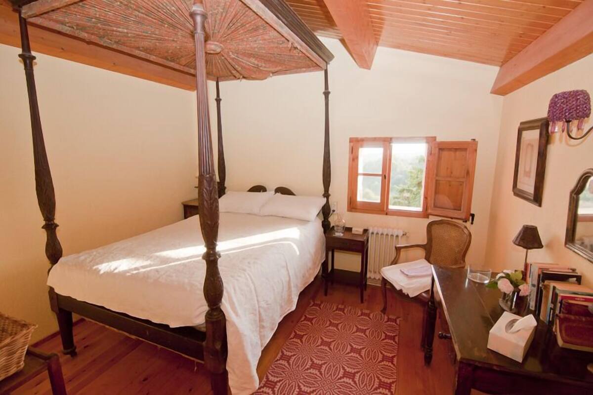 A four-poster bed with a canopy, a wooden desk with a chair, a radiator, and a window with a view of greenery.