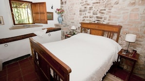 A bedroom with a wooden bed, stone walls, a window with wooden shutters, and a small bedside table with a lamp. - Celler Colltor (Santa Maria d'Oló)