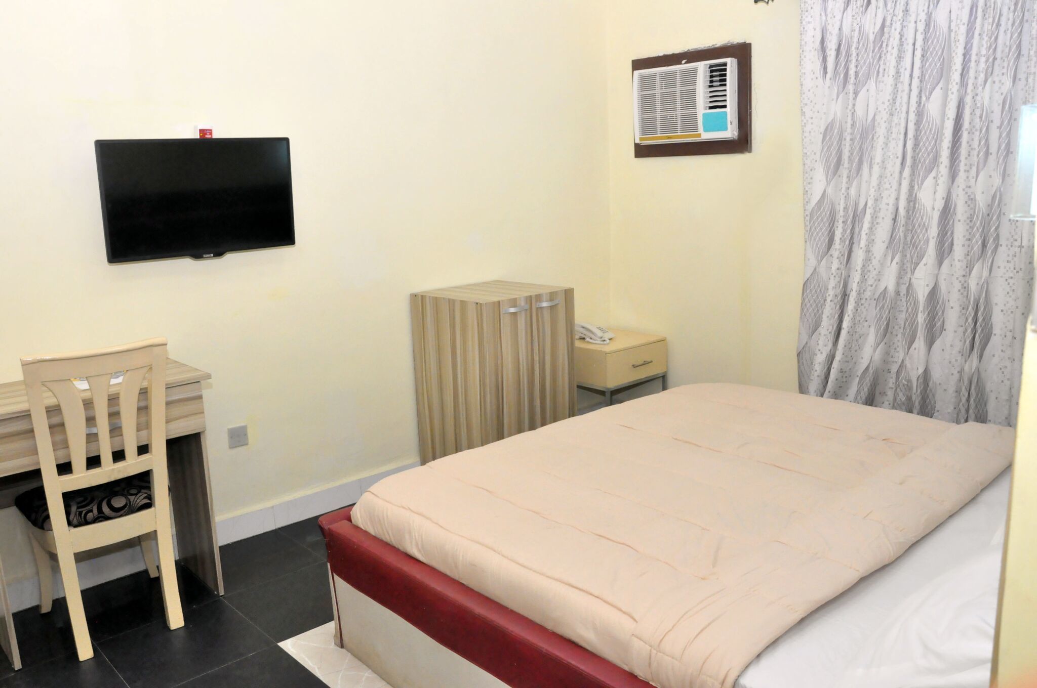 A bedroom with a bed, a desk, a chair, a wardrobe, a TV mounted on the wall, and a window with curtains.