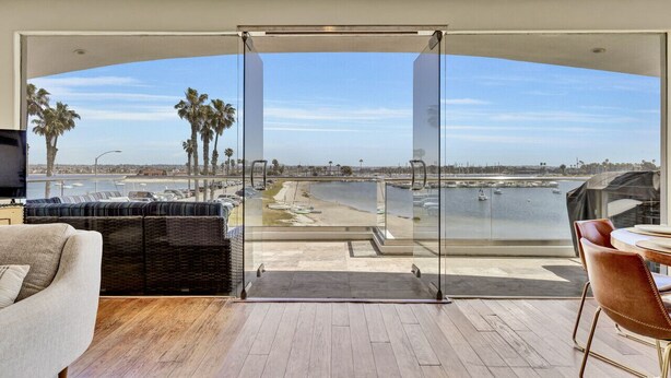Ein modernes Wohnzimmer mit Glastür, die auf einen Balkon mit Blick auf einen Hafen und Strand führt.