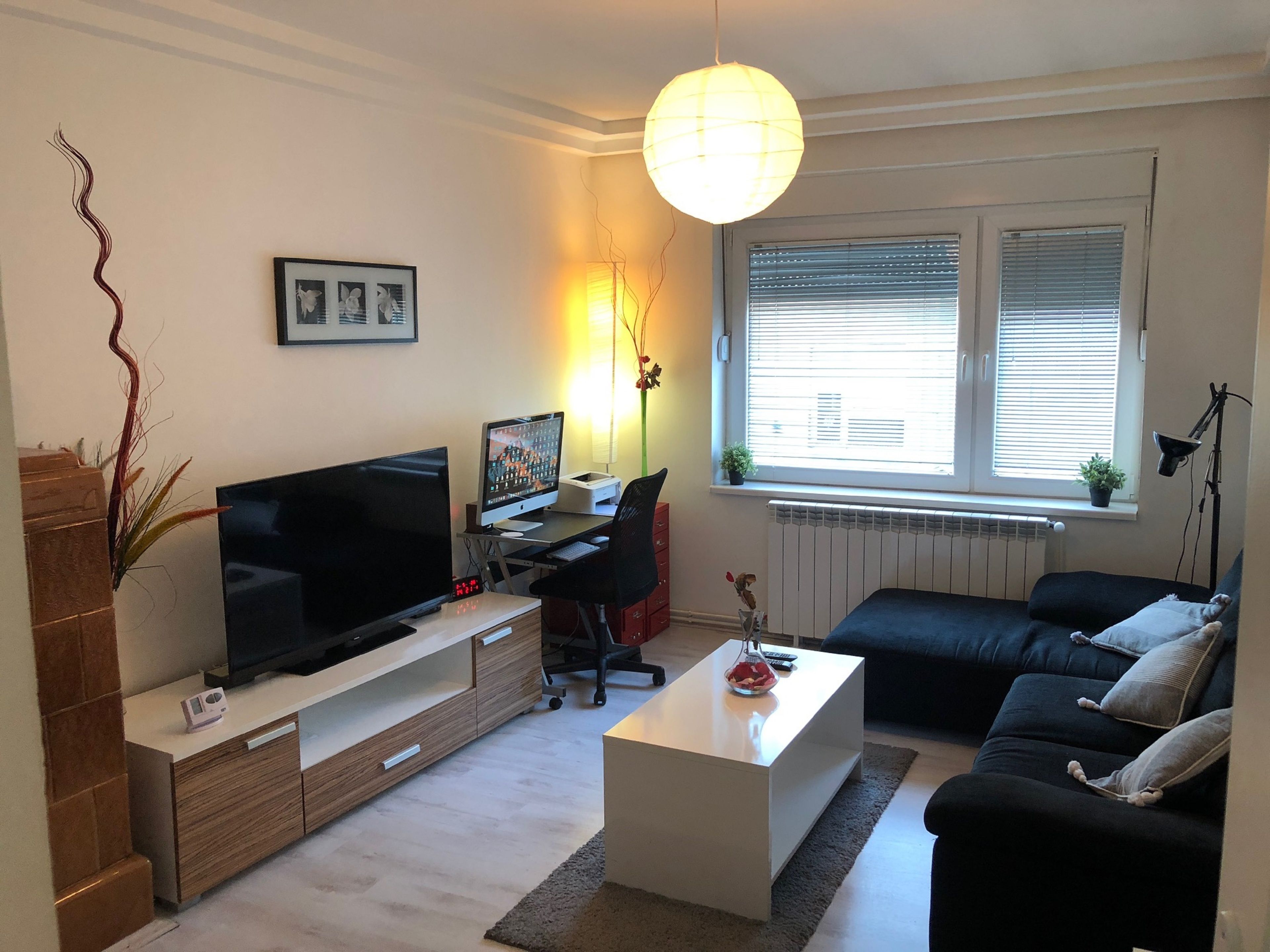 Uma sala de estar moderna com televisão de tela plana, uma escrivaninha, um sofá preto e uma mesa de centro branca.