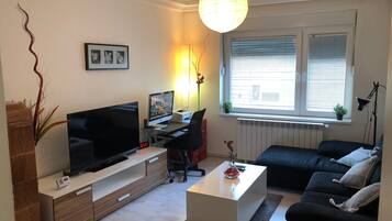 Uma sala de estar moderna com televisão de tela plana, uma escrivaninha, um sofá preto e uma mesa de centro branca.