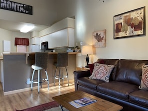 A modern living room with a brown leather sofa, two bar stools, a wooden coffee table, and a kitchen area with white cabinets and a red curtain.