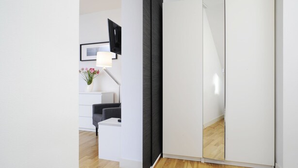 A modern room with a wooden floor, a white desk, a black chair, a white wardrobe, and a wall-mounted TV. - homefortimes Frankfurt-Airport (Langen)