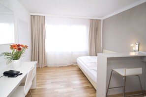 A hotel room with a bed, a desk with a telephone, a chair, a small table with a lamp, and a window with curtains. - homefortimes Frankfurt-Airport (Langen)