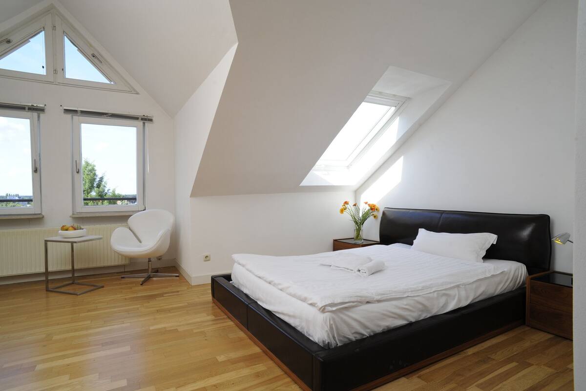 A bedroom with a bed, a chair, a small table, and a window.