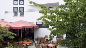 An outdoor patio with tables and chairs, a red umbrella, and a white building with multiple windows and a balcony. - homefortimes Frankfurt-Airport (Langen)