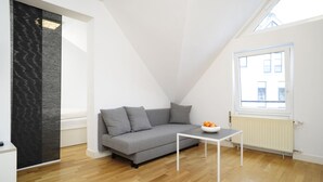 A modern living room with a grey sofa, a small white table, and a window with a view of buildings. - homefortimes Frankfurt-Airport (Langen)