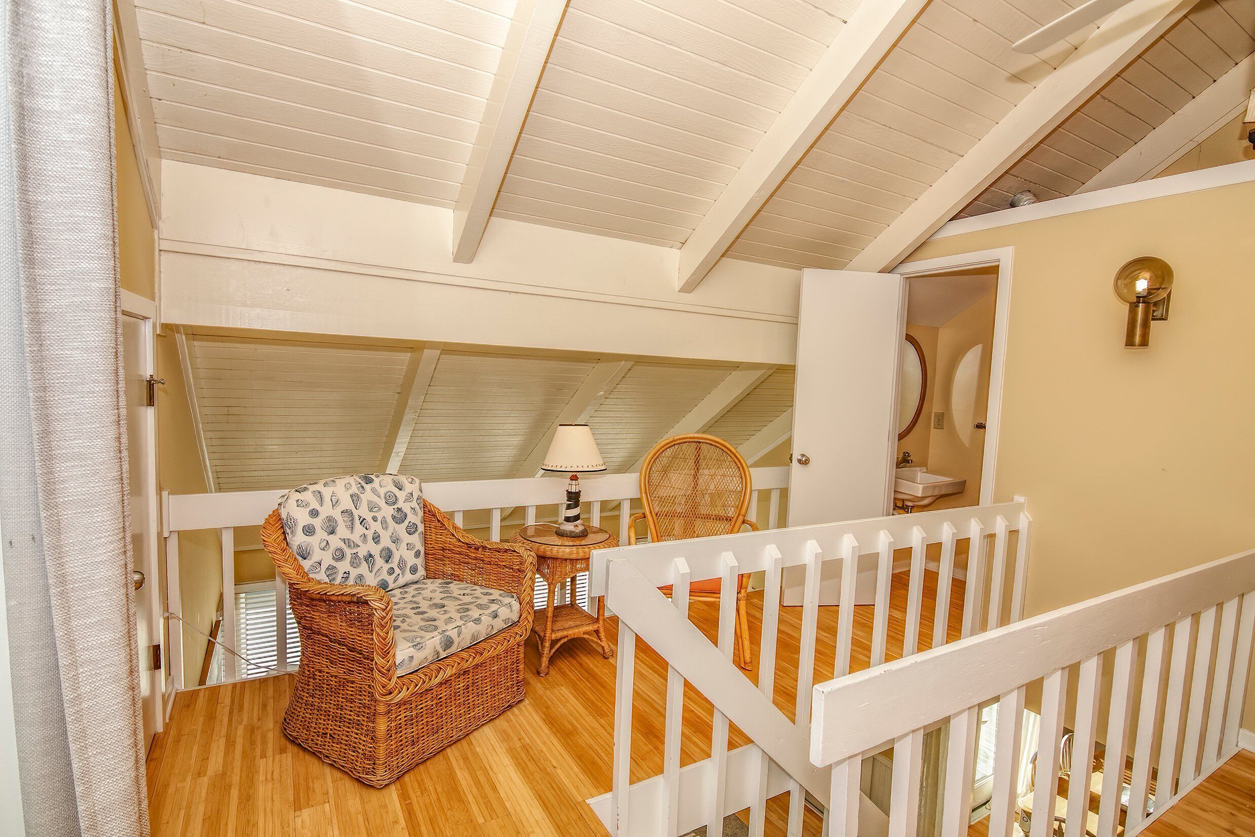 Une chambre mansardée accueillante, avec une balustrade blanche, un fauteuil en osier, une petite table avec une lampe et une vue sur la salle de bain.