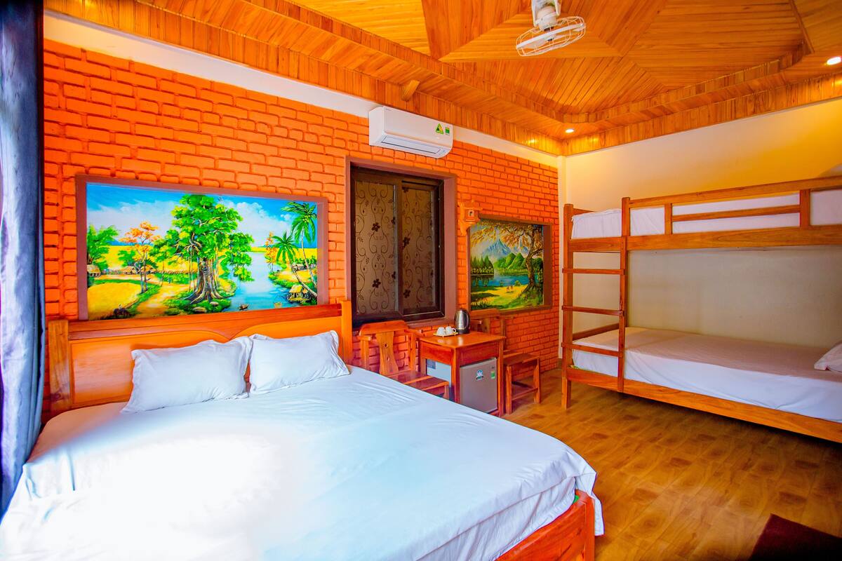 A room with a large bed, a desk, and a bunk bed. There are paintings on the wall and an air conditioner above the desk.