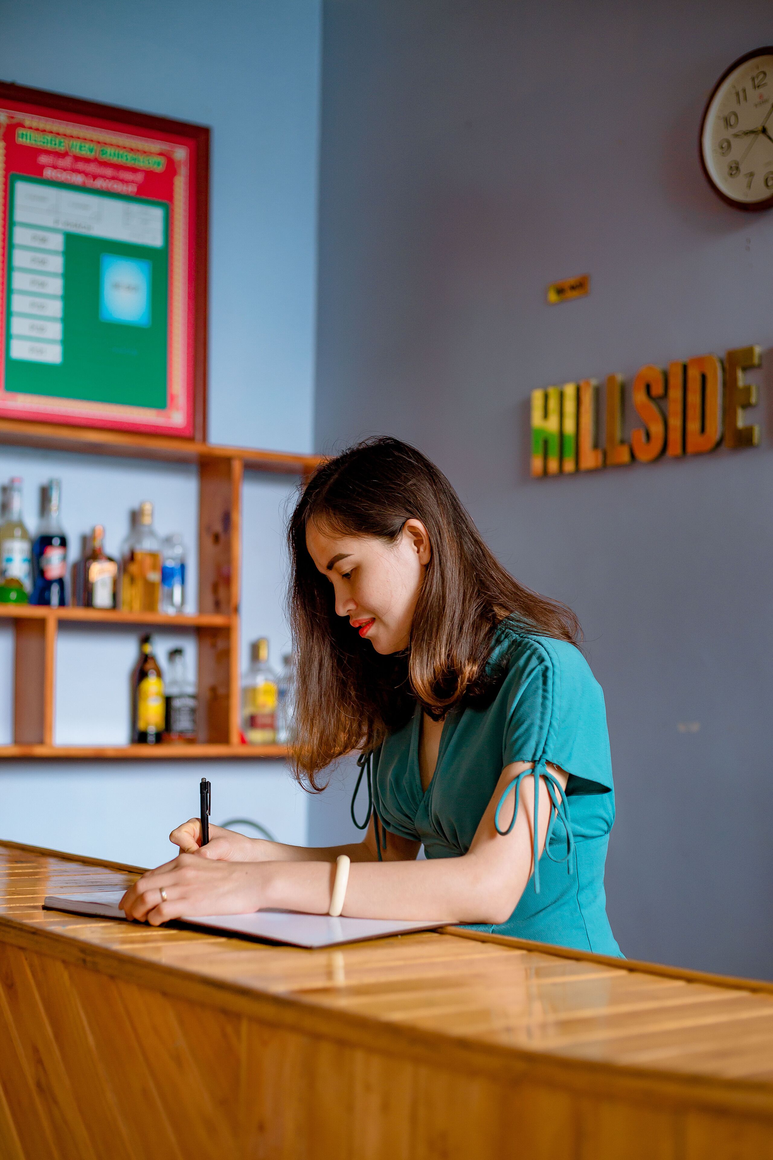 A reception desk with a person writing on a notepad. Behind the desk, there is a bulletin board with a red sign and a clock on the wall.