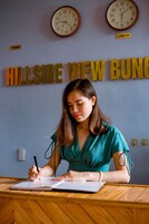 Una persona escribiendo en un escritorio de madera con un cuaderno, frente a una pared con relojes y el texto "HILLSIDE VIEW BUNG".