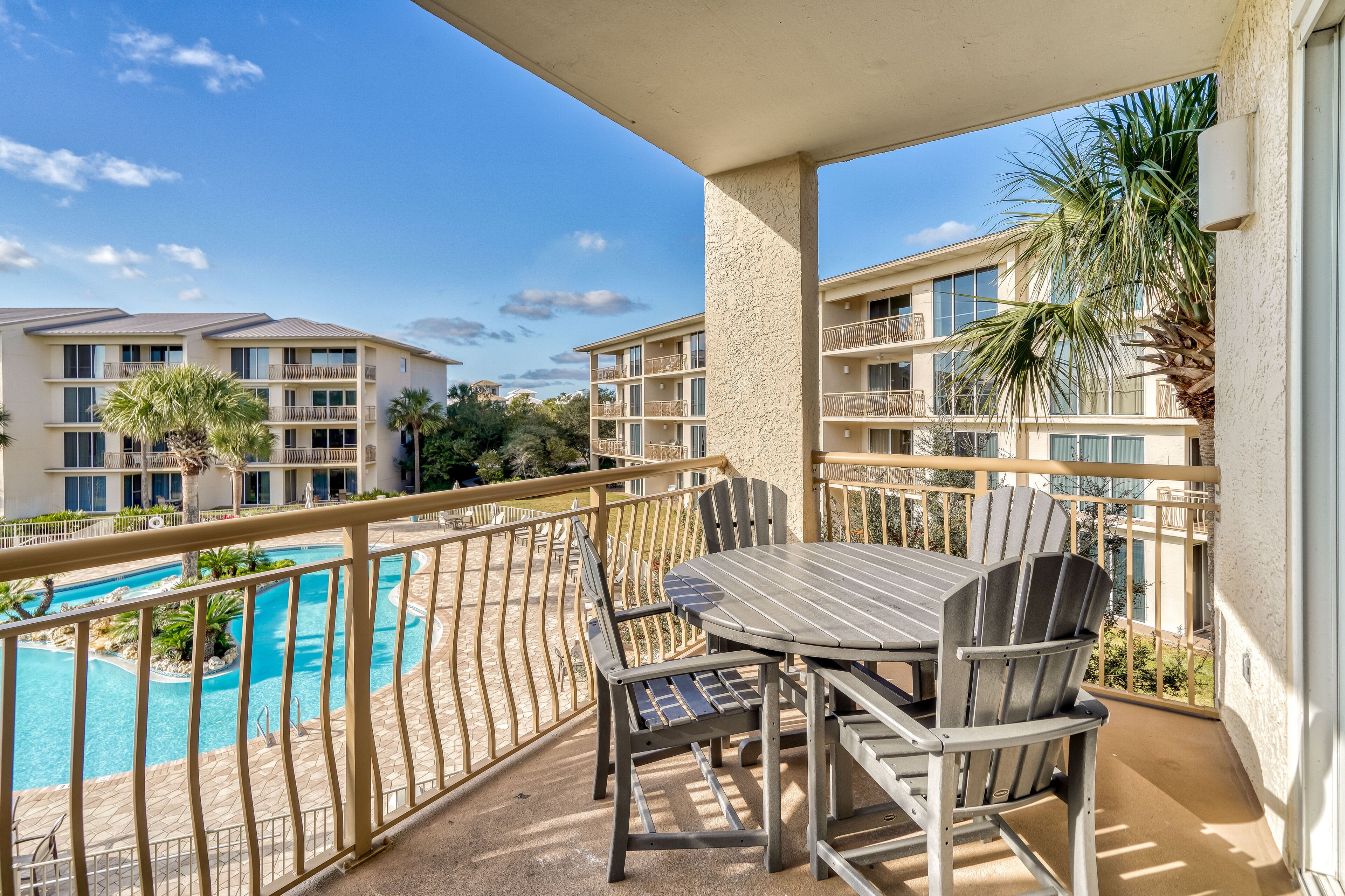 A balcony with a table and chairs overlooking a swimming pool, surrounded by a fence and palm trees.