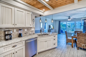 A kitchen with white cabinets, a granite countertop, a dishwasher, and a dining area with wicker chairs. - HP 315 Condo @ High Pointe | Lagoon & Kids’ Pool | Tennis & Pickleball Courts (High Pointe Resort)