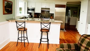 A modern kitchen with white cabinets, stainless steel appliances, and a wooden floor. There are two bar stools and a plaid chair. - Classic and Spacious Maine Waterfront Retreat (Sullivan)