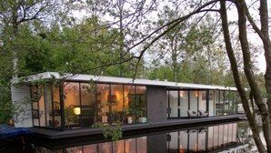 A modern houseboat with large glass windows, a flat roof, and a water body in front.
