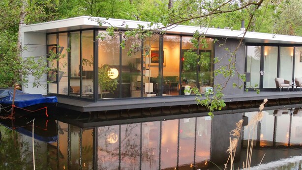 A modern houseboat with large glass windows, a deck with seating, and a view of the surrounding water and greenery.