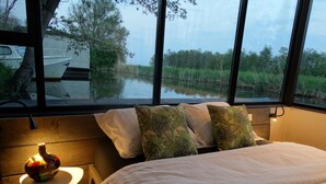 A bedroom with a large window, a bed with white bedding, two pillows with leaf patterns, and a small table with a lamp.