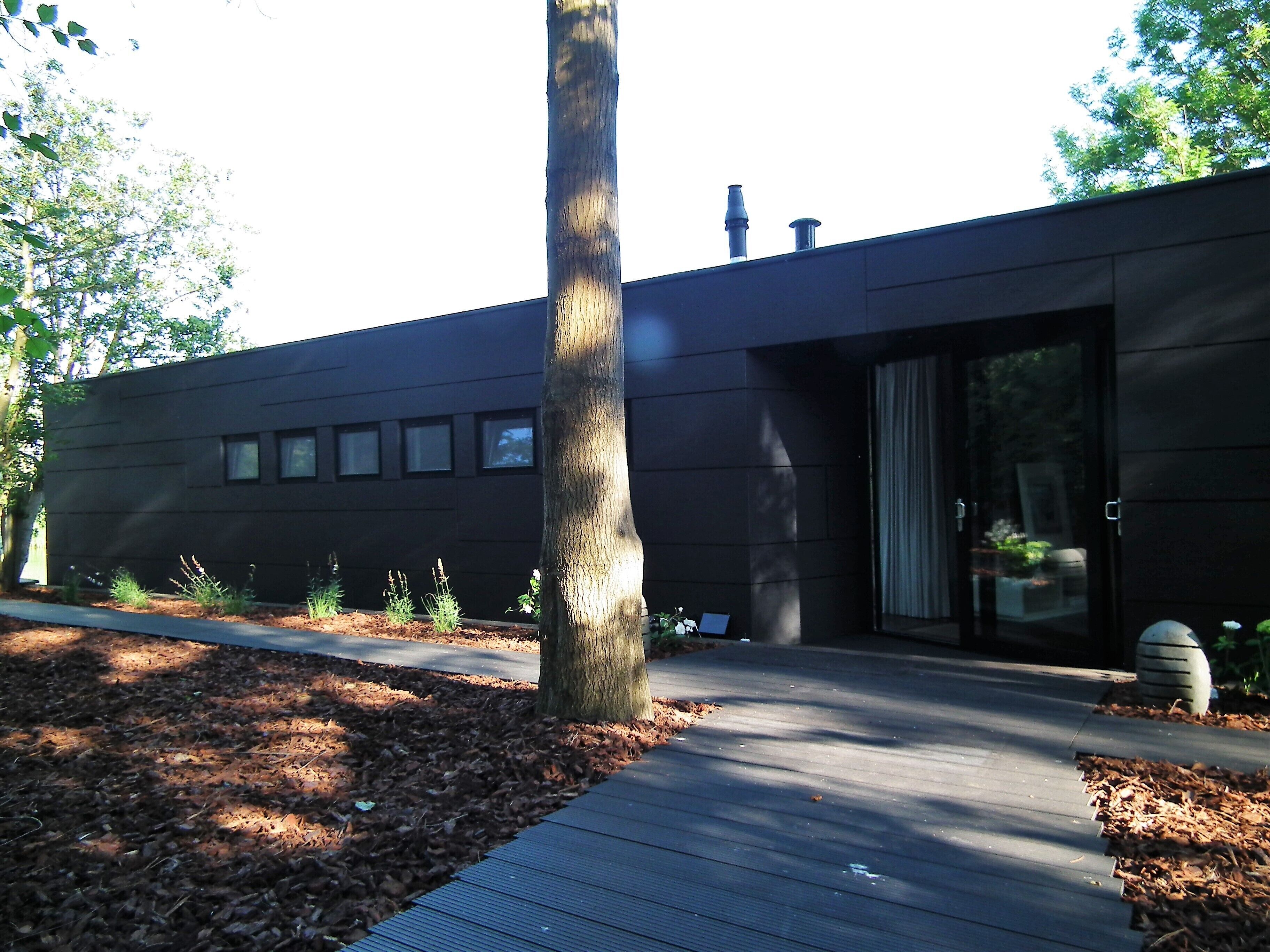 Ein modernes Gebäude mit dunkler Fassade, einer Holzterrasse und einem Weg zu einer Glastür.
