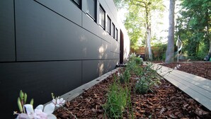 A modern building with a dark facade, a wooden deck, and a garden area with mulch and plants.