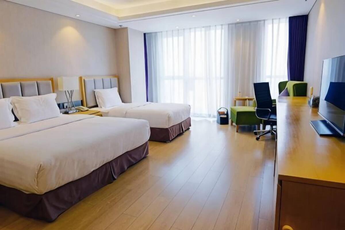 A hotel room with two beds, a desk, a chair, a TV, and a large window with curtains.