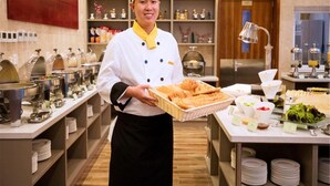 A chef in a professional kitchen holding a basket of pastries. The kitchen is equipped with stainless steel appliances and has a variety of food items and condiments on display. - Ariva Tianjin Zhongbei Serviced Apartment (Tianjin)