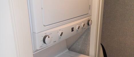 A compact laundry room with a front-loading washer and dryer, a control panel with various settings, and a small trash bin.