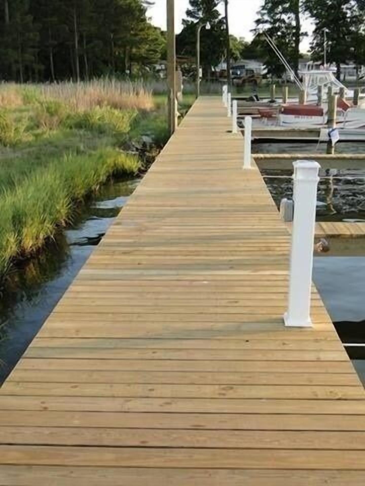 Une promenade en bois menant à un quai où sont amarrés des bateaux, le tout étant entouré de zones herbeuses et d’arbres.
