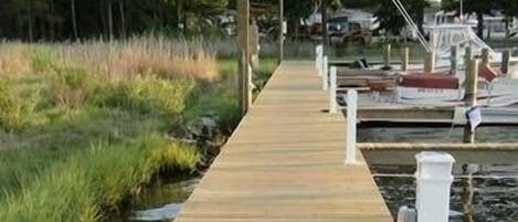 Une promenade en bois menant à un quai où sont amarrés des bateaux, le tout étant entouré de zones herbeuses et d’arbres.