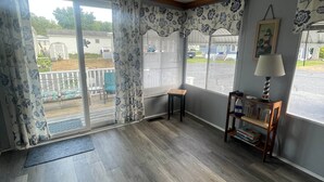 Ein Zimmer mit einer großen Schiebeglasdoor, die auf eine Terrasse führt, einem kleinen Holztisch, einer Lampe und einem Bücherregal.