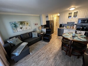 Un salon avec un canapé foncé, une table basse et un coin repas avec des chaises. Au fond, on aperçoit une cuisine avec des meubles blancs et des appareils électroménagers.