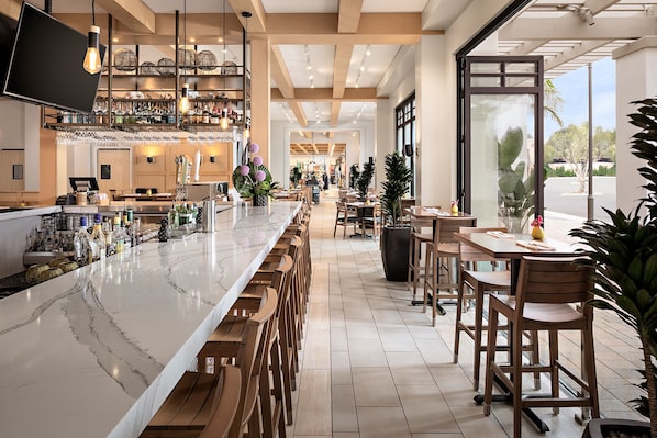 A modern bar area with a long counter, wooden bar stools, and a variety of bottles and glasses.