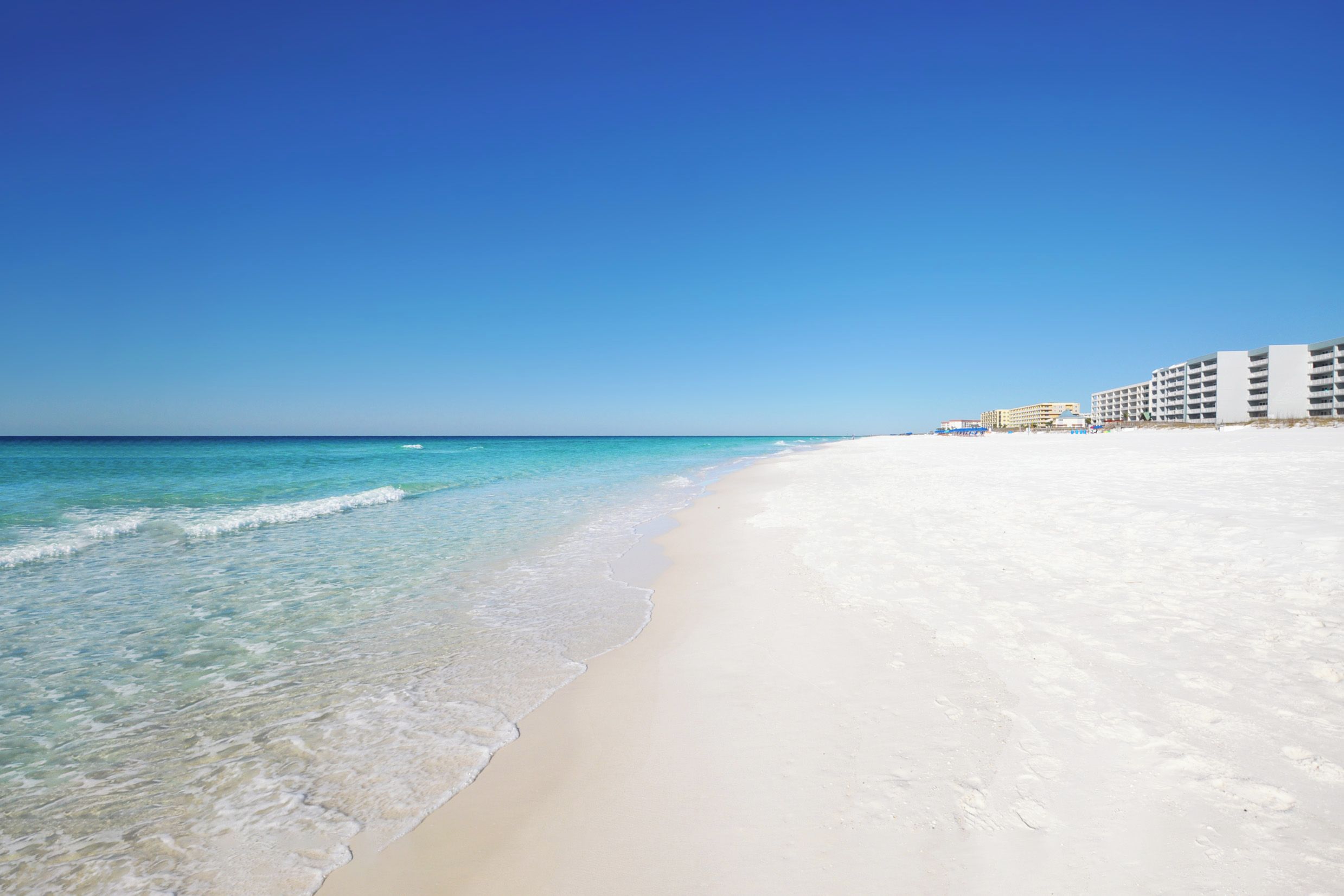 A beach with clear water, a sandy shore, and a hotel with multiple floors in the background.