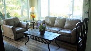 A room with a leather sofa, a matching armchair, a small table with a lamp and flowers, and large windows with a view of greenery.