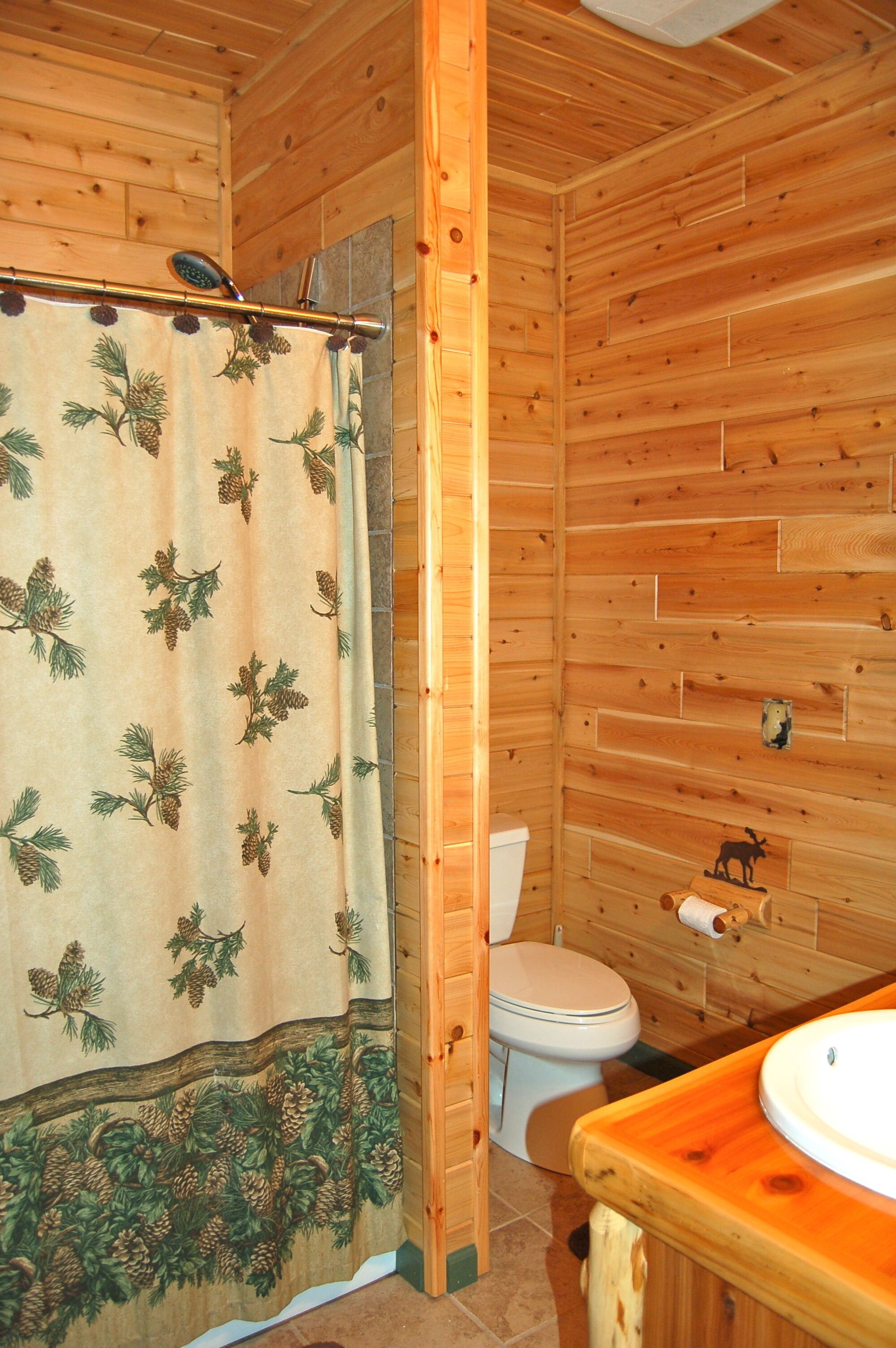 A wooden cabin bathroom with a pine leaf shower curtain, a toilet, and a wooden sink.