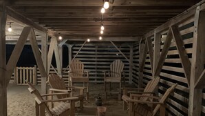 Et overdækket udendørs opholdsområde med træstole og -borde, sandgulv og et træhegn i baggrunden.