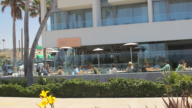 An outdoor dining area with tables and umbrellas, surrounded by palm trees and a building with large windows. - Chic Beach Apartment..Steps to the sand! (San Diego)